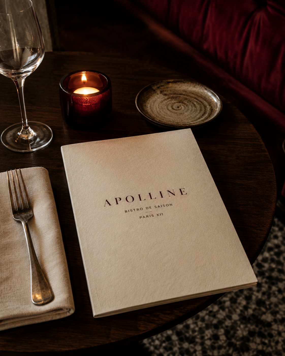 Table ronde en bois sombre avec la carte fermée d'Apolline, bougie, assiette en céramique et banquette lie-de-vin.